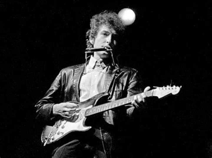 Bob Dylan plays a guitar and Mick Jagger claps his hands. They both stand in front of microphones.