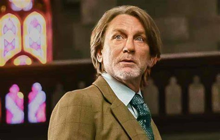 Daniel Craig as Benoit Blanc for our article about 'Knives Out 3.' He's wearing a blue collared shirt, beige jacket, and leaning on a stand.