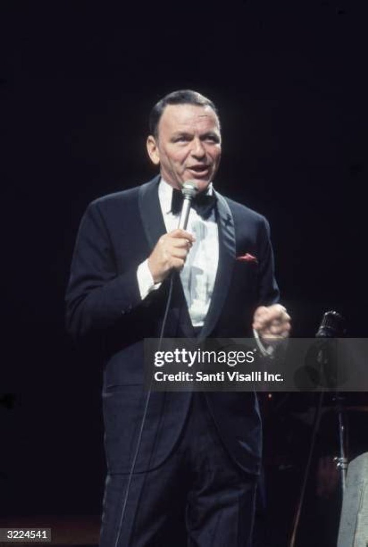 Frank Sinatra wears a tuxedo and stands in front of a microphone.