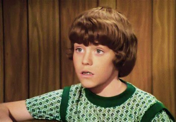 Mike Lookinland as Bobby Brady, wearing a baseball hat, and in 2019