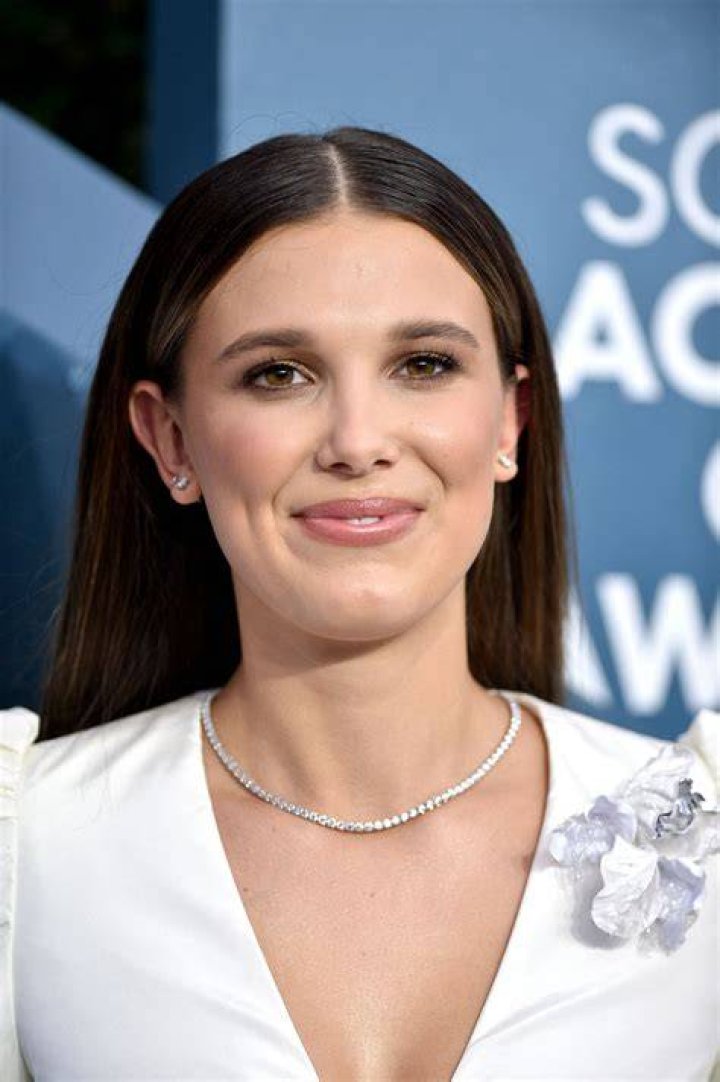 Millie Bobby Brown at the 'Stranger Things 3' premiere wearing a blue dress with flowers in front of the 'Stranger Things 3' promo poster.